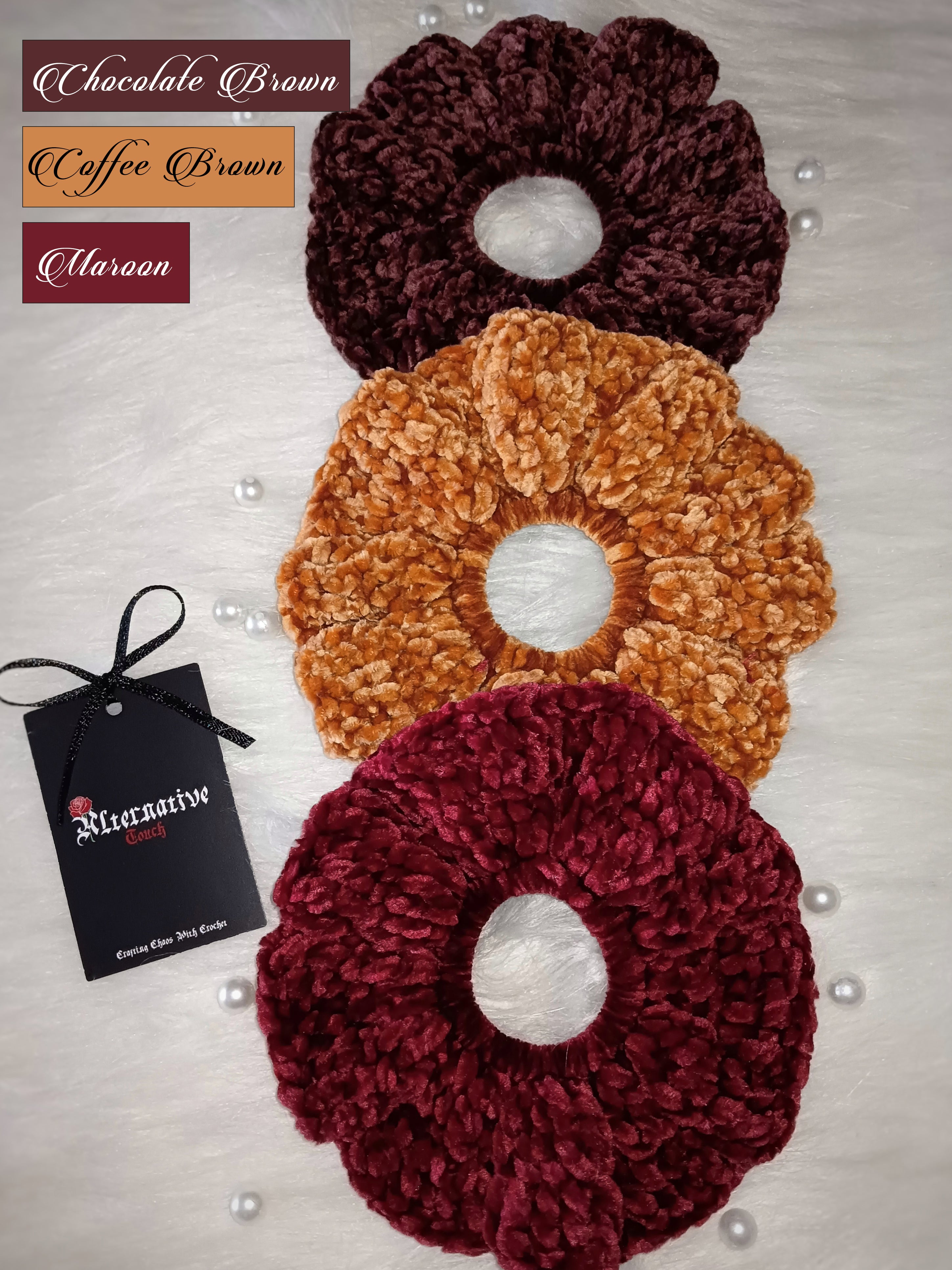 Three crocheted velvet hair scrunchies in chocolate brown, coffee brown, and maroon with a brand tag on a light background.