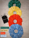 Four colorful scrunchies in the shades peacock green, light yellow, orange and sky blue on a light surface with a brand tag and color swatches below.