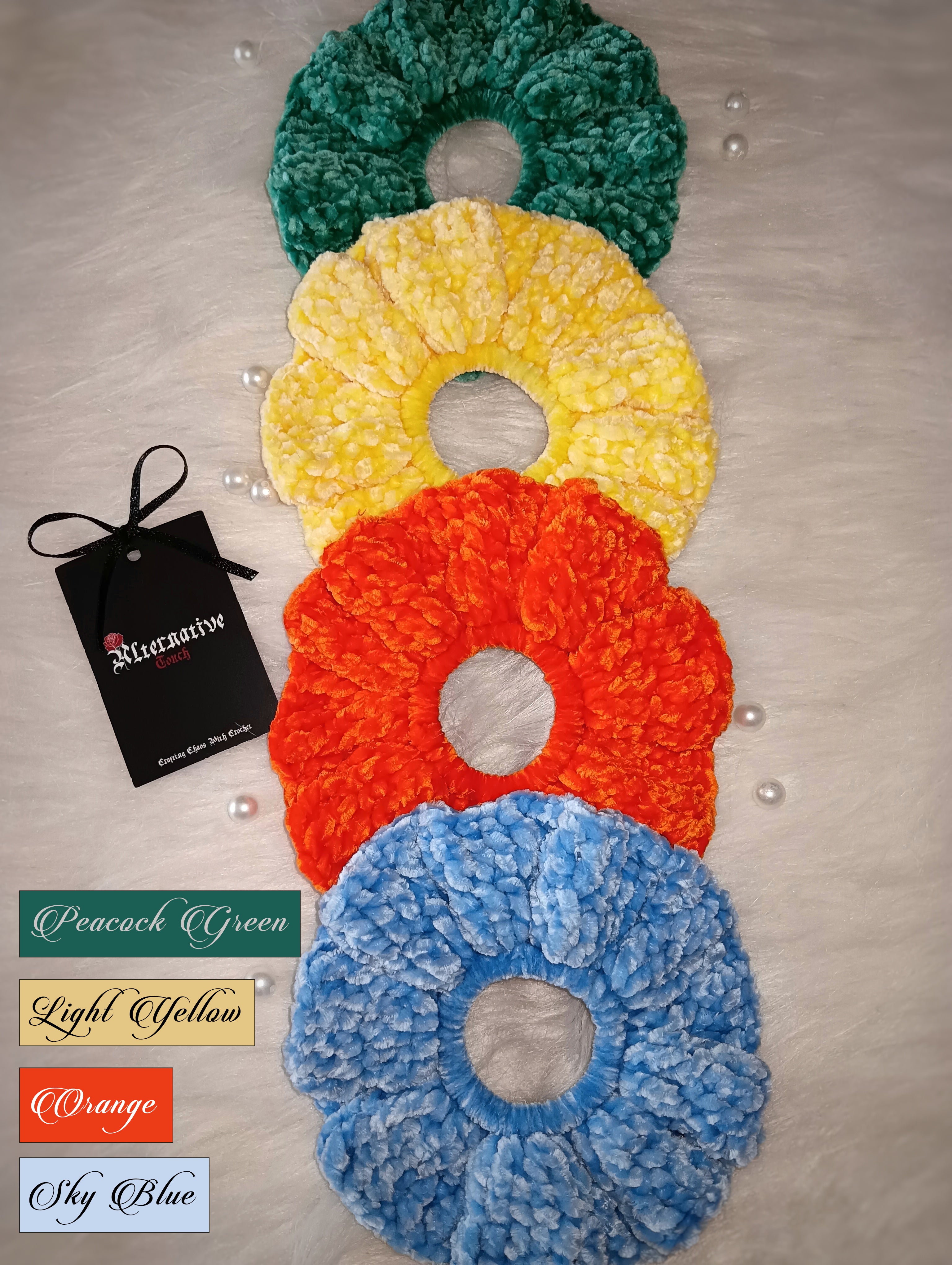 Four colorful scrunchies in the shades peacock green, light yellow, orange and sky blue on a light surface with a brand tag and color swatches below.